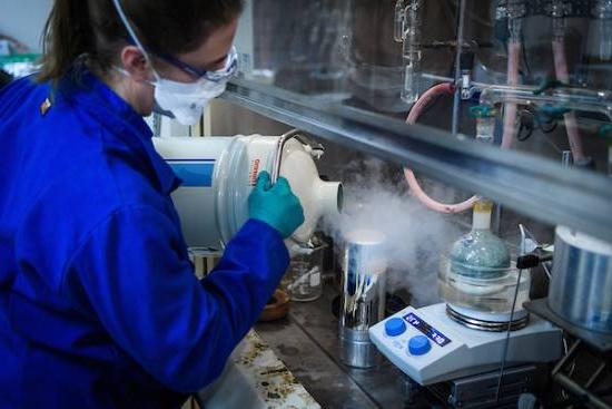 person in lab coat and gear pours big vessel of steaming substance into smaller container under fume hood