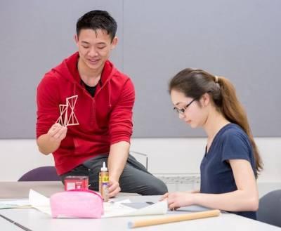 students work together.  One sits at table, one sits on table.  they work with wood pieces and glue.